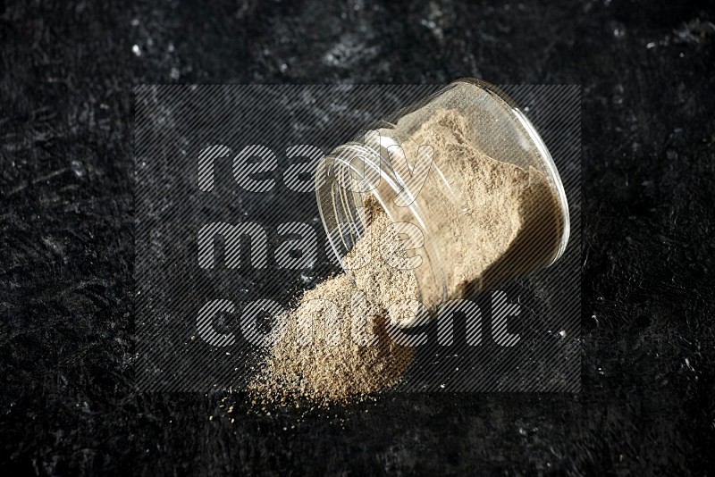 A flipped glass jar full of cardamom powder and powder spilled out of it on textured black flooring