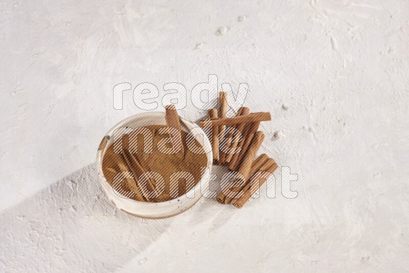 Ceramic bowl full of cinnamon powder with cinnamon sticks on the side on white background