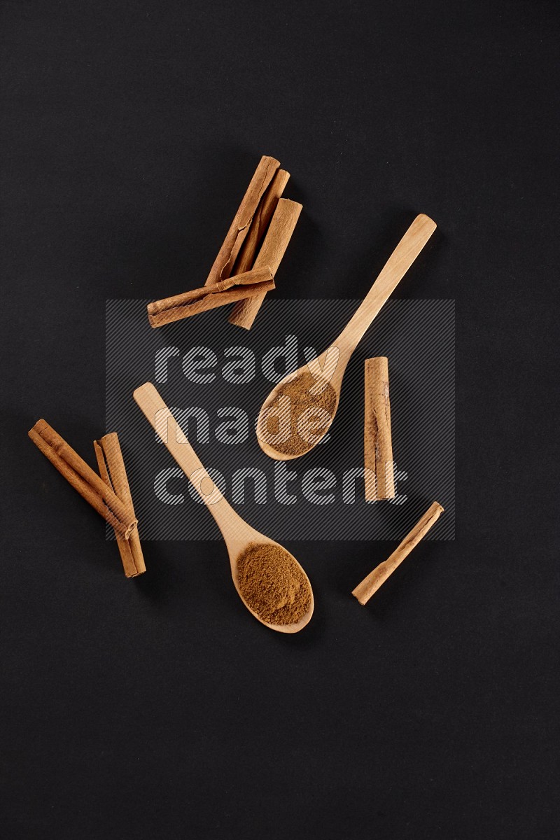 Cinnamon powder in two wooden spoons with cinnamon sticks on black background