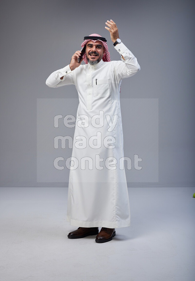 Saudi man Wearing Thob and red Shomag standing talking on phone on Gray background