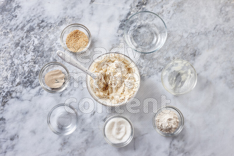 كحك خطوة بخطوة بمكوناته سمن، سكر، دقيق، سمسم، خميرة، ماء ونكهة الكحك على خلفية رخام رمادي