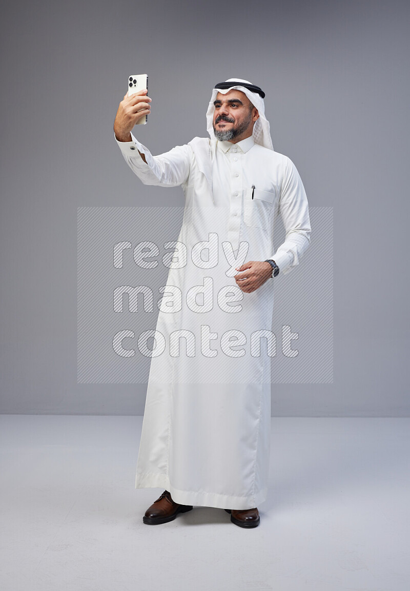 Saudi man Wearing Thob and white Shomag standing taking selfie on Gray background