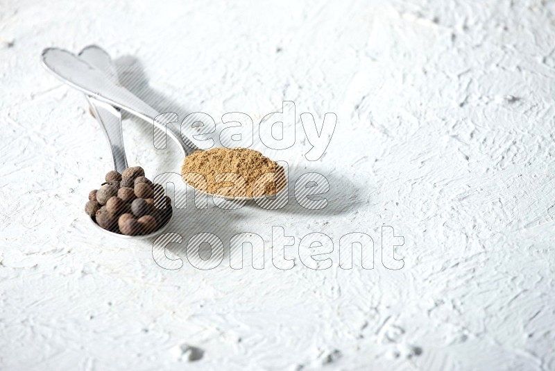 2 metal spoons full of allspice powder and whole balls on a textured white flooring