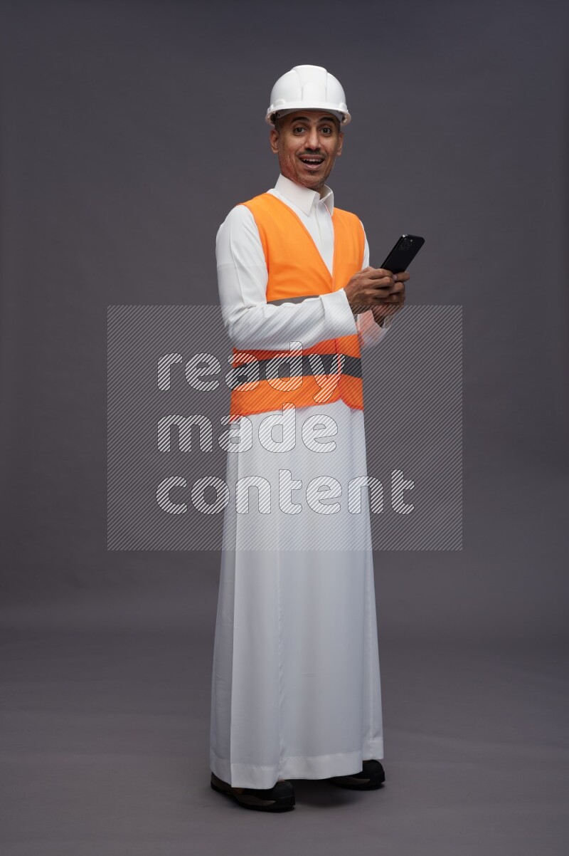 Saudi man wearing thob with engineer vest standing texting on phone on gray background