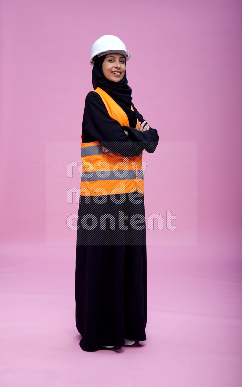 Saudi woman wearing Abaya with engineer vest and helmet standing with crossed arms on pink background