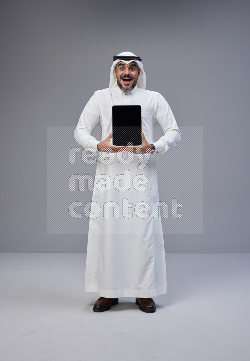 Saudi man Wearing Thob and white Shomag standing showing tablet to camera on Gray background