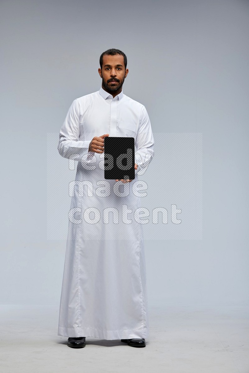 Saudi man wearing Thob standing showing tablet to camera on Gray background