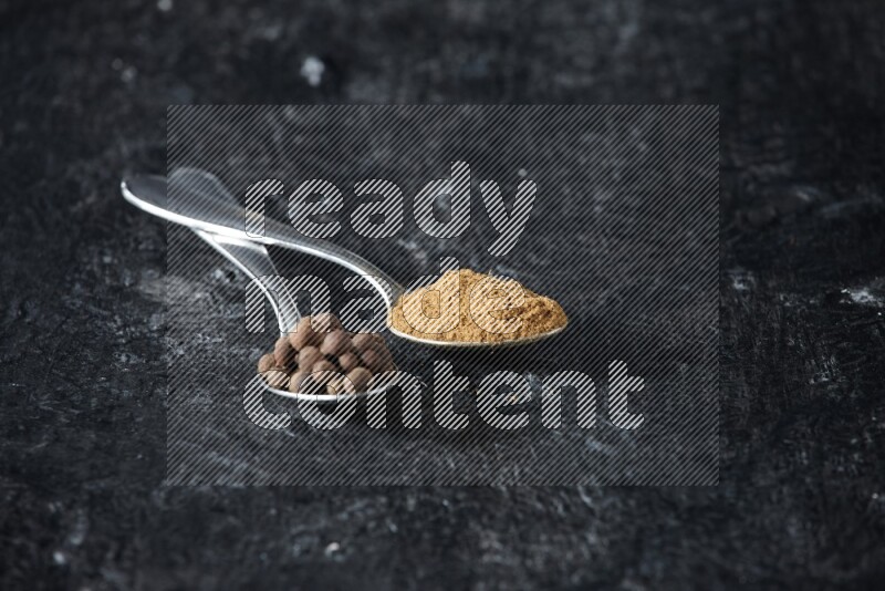 ملعقتان معدنيتان ممتلئتان ببودرة بهار الفلفل الحلو وحبوبه على خلفية سوداء