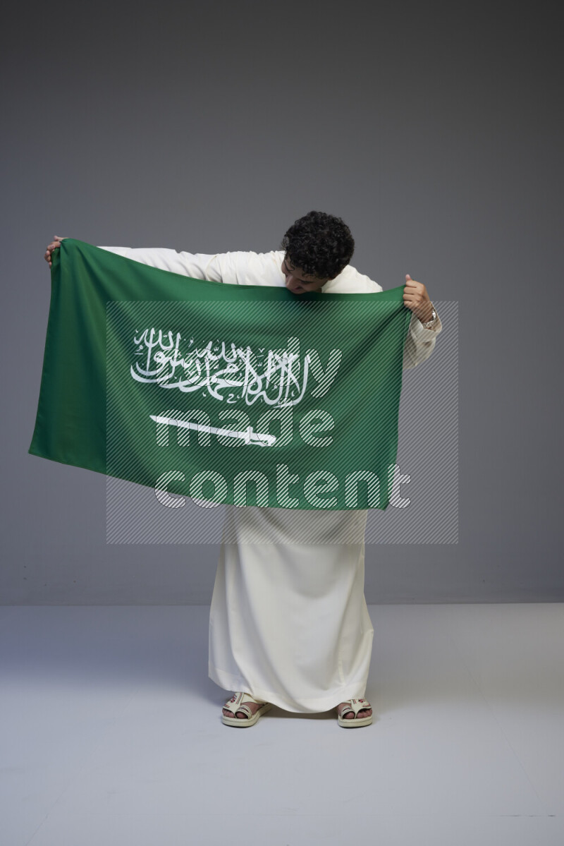 A Saudi man standing wearing thob holding big Saudi flag on gray background