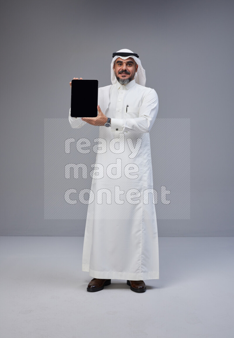 Saudi man Wearing Thob and white Shomag standing showing tablet to camera on Gray background