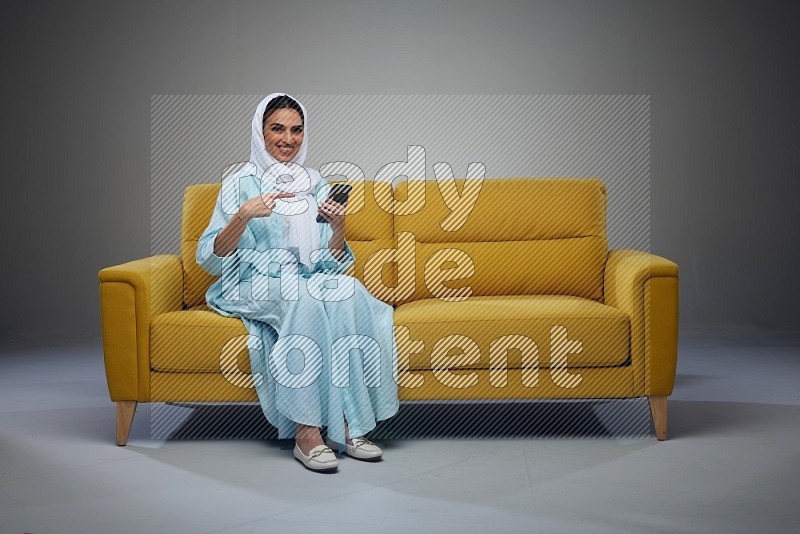 A Saudi woman wearing a light blue Abaya and a white head scarf sitting on a yellow sofa and holding her phone while pointing to it eye level on a grey background
