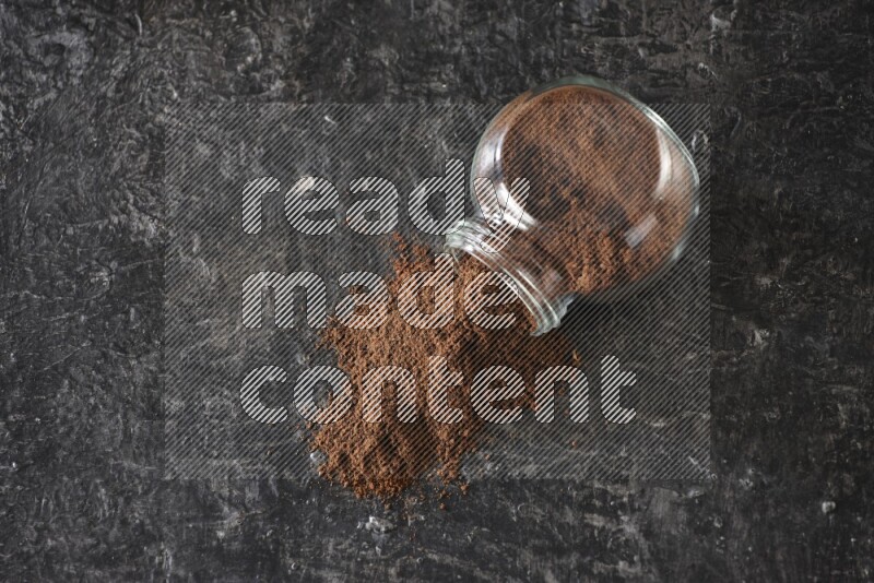 A flipped glass spice jar full of cloves powder and powder came out of it on textured black flooring