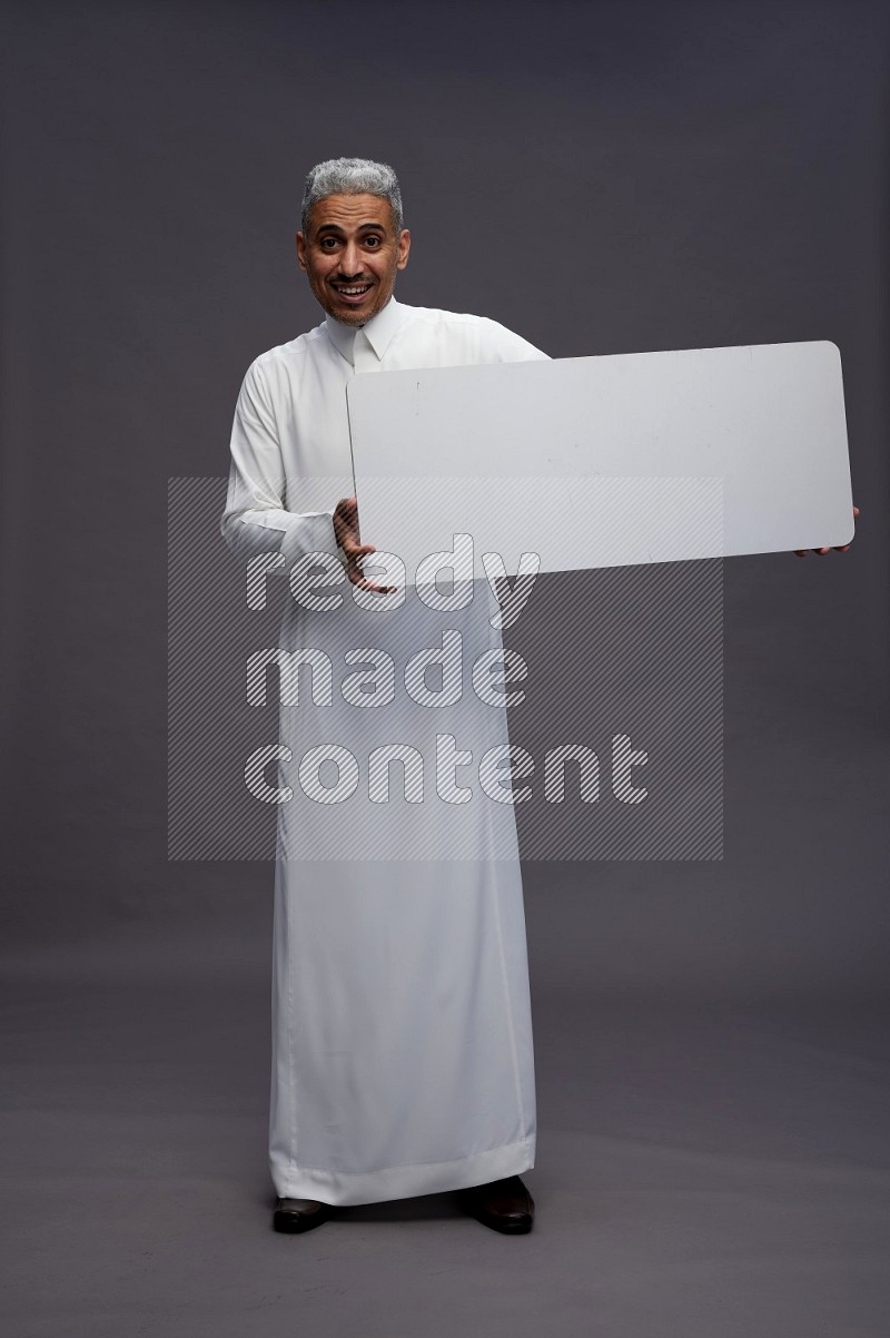 Saudi man wearing thob standing holding board on gray background