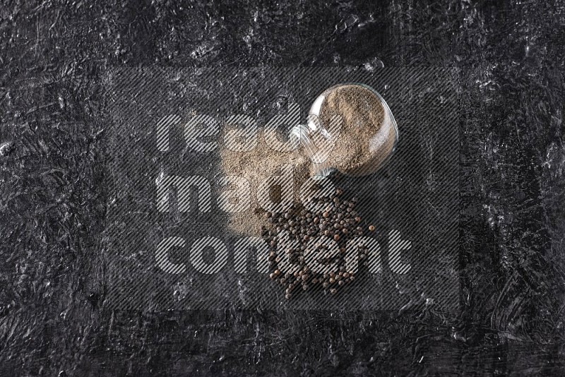 A flipped glass spice jar full of black pepper powder with pepper beads on textured black flooring