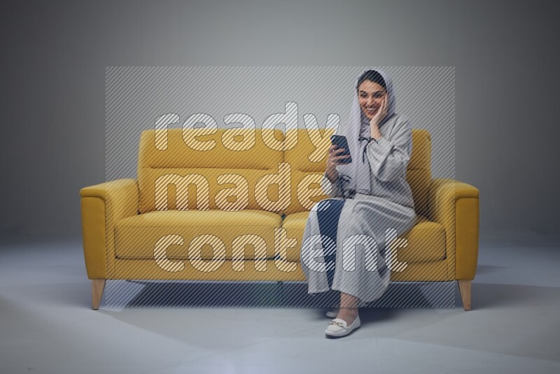 A Saudi woman wearing a light gray Abaya and white head scarf sitting on a yellow sofa and using her phone eye level on a grey background