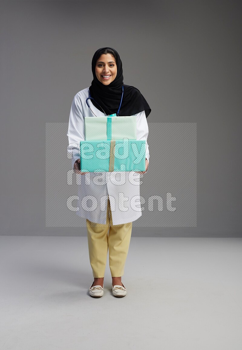 Saudi woman wearing lab coat with stethoscope standing holding gift box on Gray background