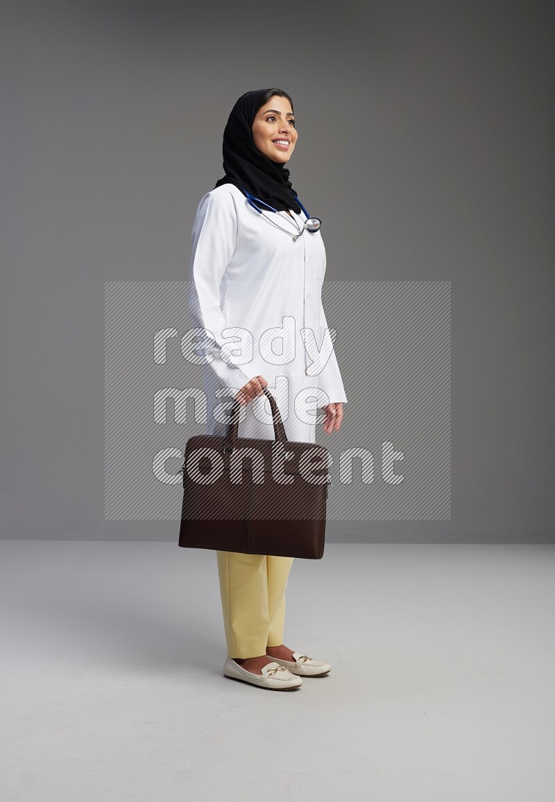 Saudi woman wearing lab coat with stethoscope standing holding bag on Gray background
