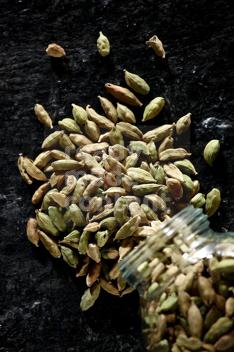 A flipped glass spice jar full of cardamom seeds on textured black flooring