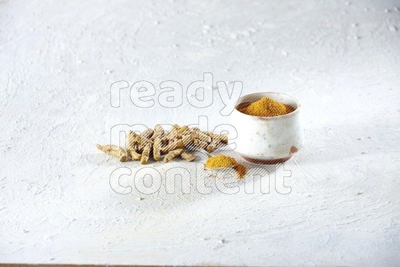 A beige pottery bowl and metal spoon full of turmeric powder and dried turmeric fingers next of them on textured white flooring