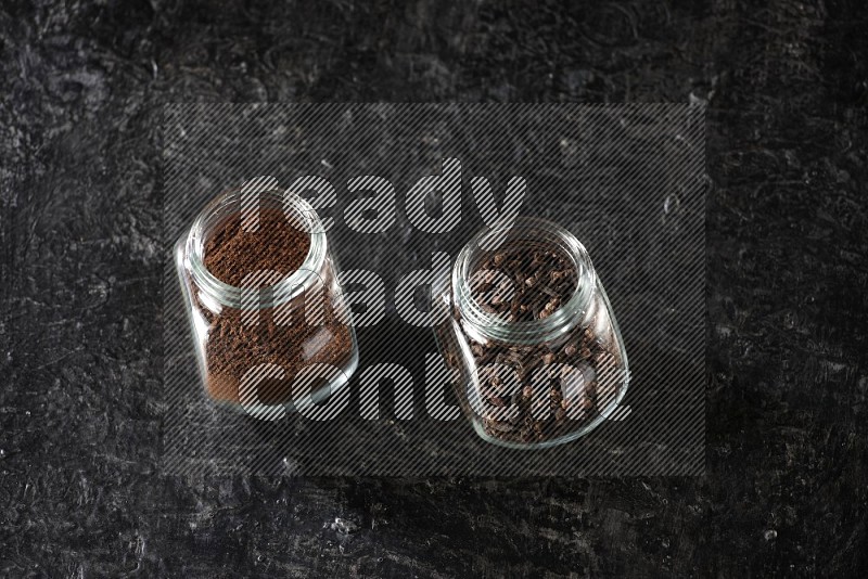 Glass spice jars full of cloves and cloves powder on textured black flooring