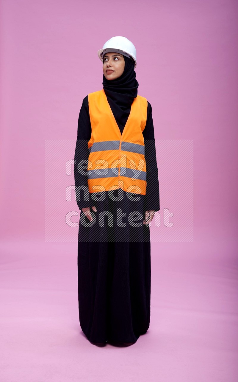 Saudi woman wearing Abaya with engineer vest and helmet standing interacting with the camera on pink background