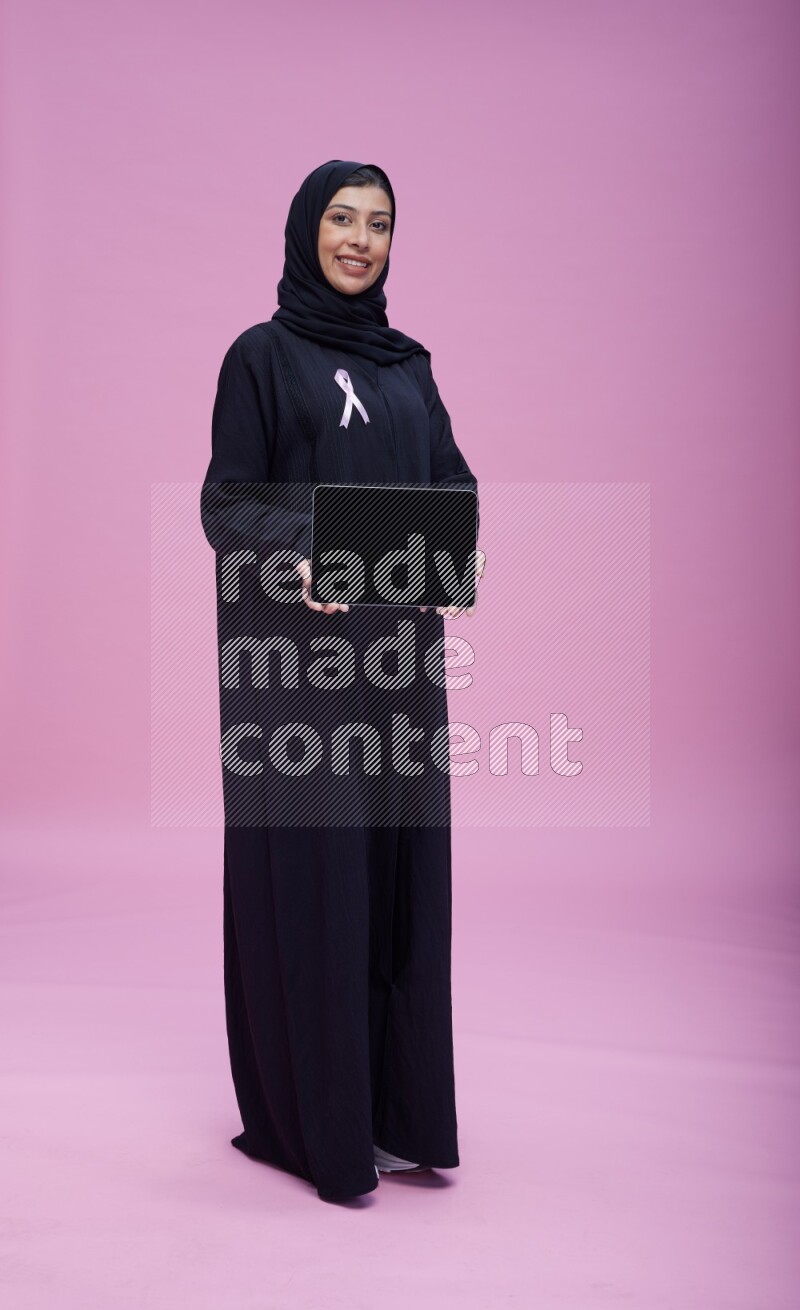 Saudi woman wearing pink ribbon on Abaya standing showing a tablet to camera on pink background