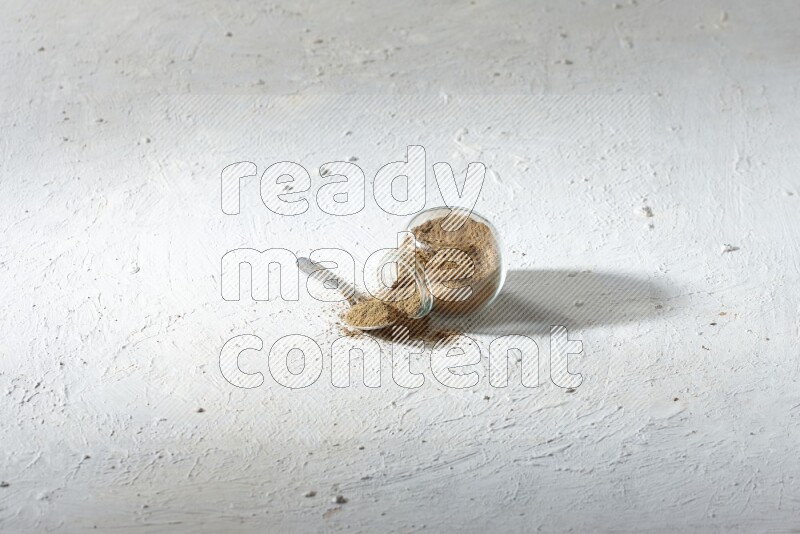 وعاء زجاجي للتوابل مقلوب وملعقة معدنية كلاهما ممتلئ ببودرة الكمون والبودرة تناثرت منه على خلفية بيضاء