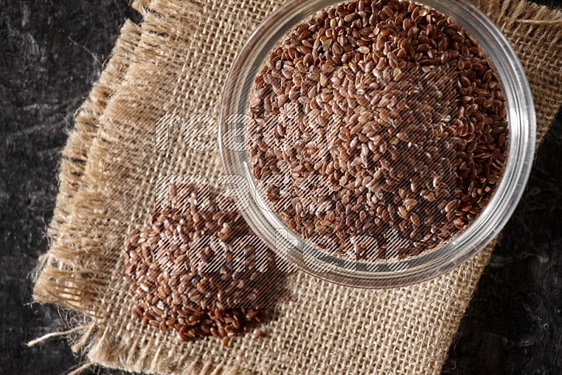 A glass bowl full of flaxseeds with bunch of the seeds on burlap fabric on a textured black flooring