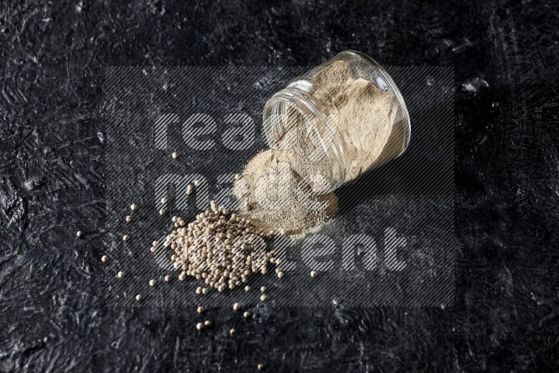 A flipped glass jar full of white pepper powder with spilled powder and pepper beads on textured black flooring