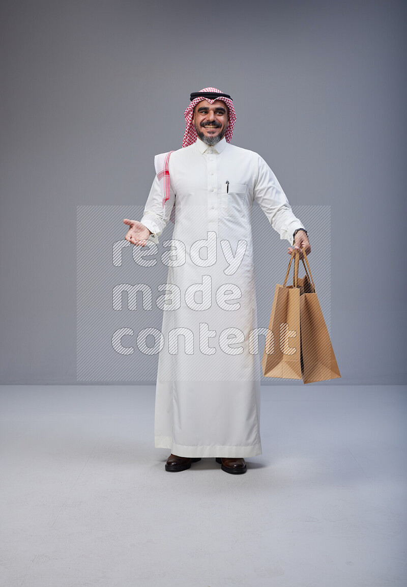 Saudi man Wearing Thob and red Shomag standing holding shopping bag on Gray background