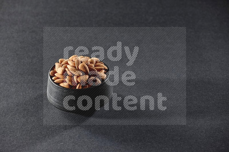 A black ceramic bowl full of peeled almonds on a black background in different angles