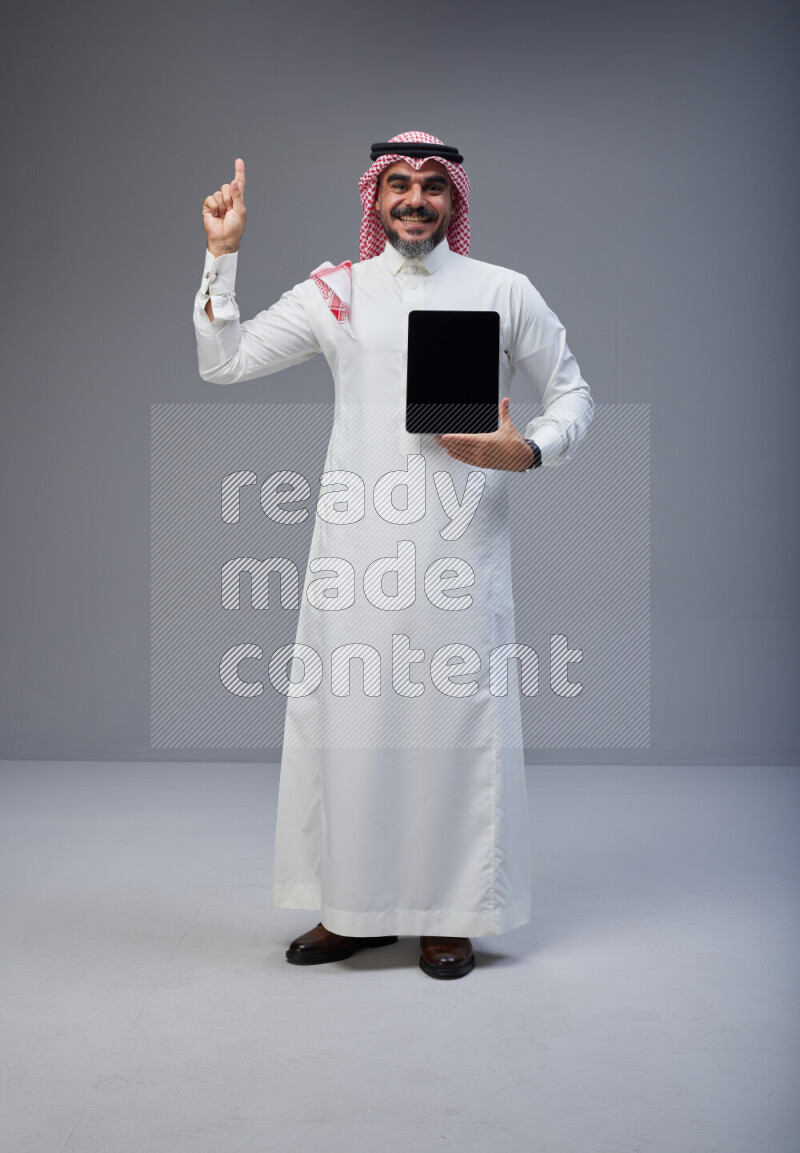 Saudi man Wearing Thob and red Shomag standing showing tablet to camera on Gray background