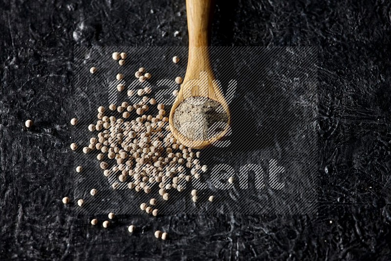 A wooden spoon full of white pepper powder with white pepper beads on textured black flooring