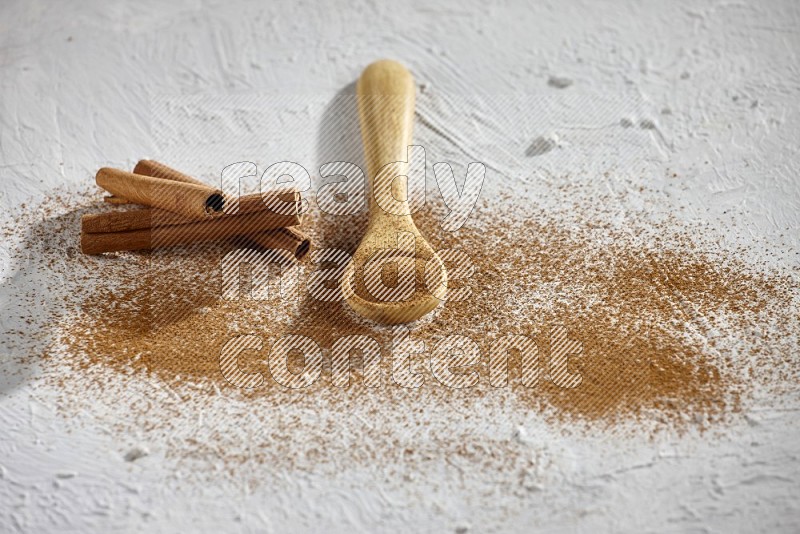 بودرة القرفة في ملعقة خشبية مع أعواد القرفة وبودرة متناثرة على الأرضية على خلفية بيضاء