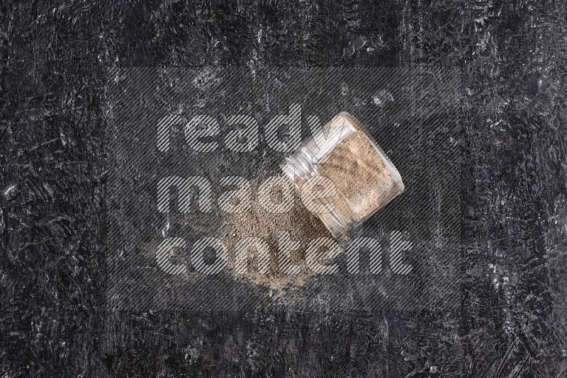 A flipped glass jar full of black pepper powder on a textured black flooring