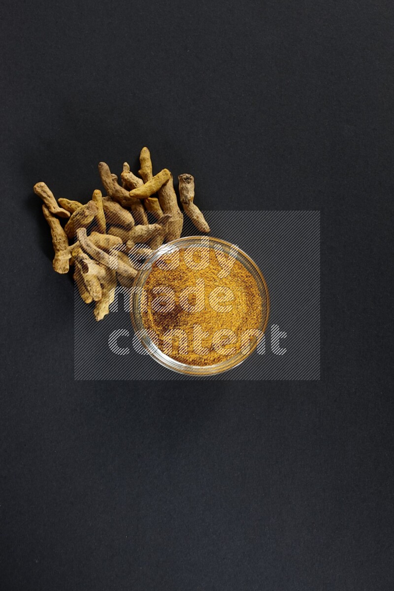 وعاء زجاجي ممتلئ ببودرة الكركم وأصابع الكركم المجففة بجانبه على خلفية سوداء