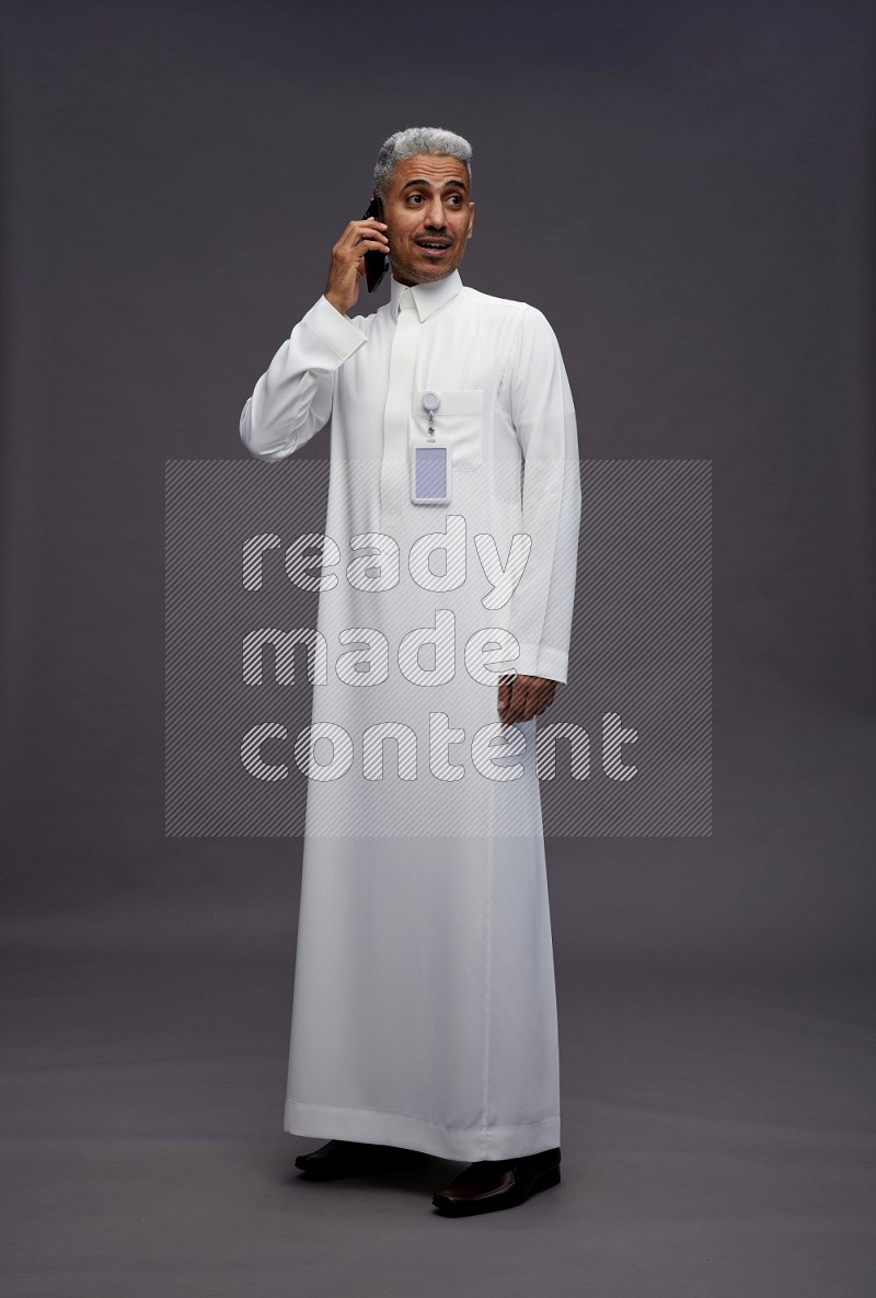 Saudi man wearing thob with pocket employee badge standing talking on phone on gray background