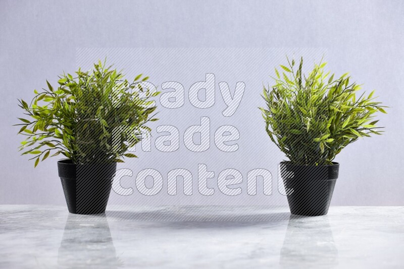 Two green artificial plants in black pots on grey marble background