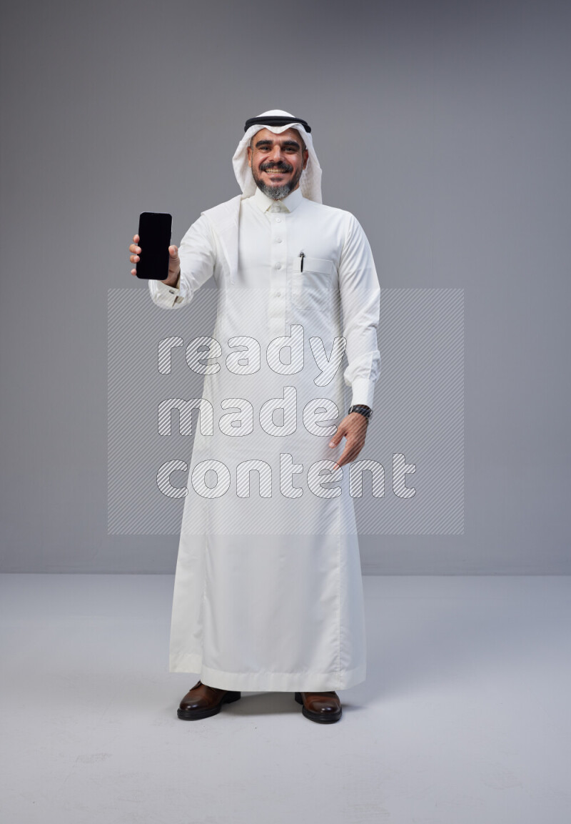 Saudi man Wearing Thob and white Shomag standing showing phone to camera on Gray background