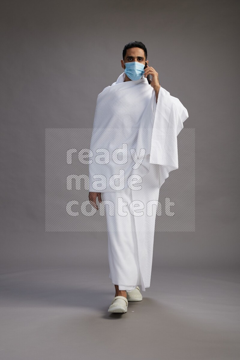A man wearing Ehram with face mask Standing talking on phone on gray background