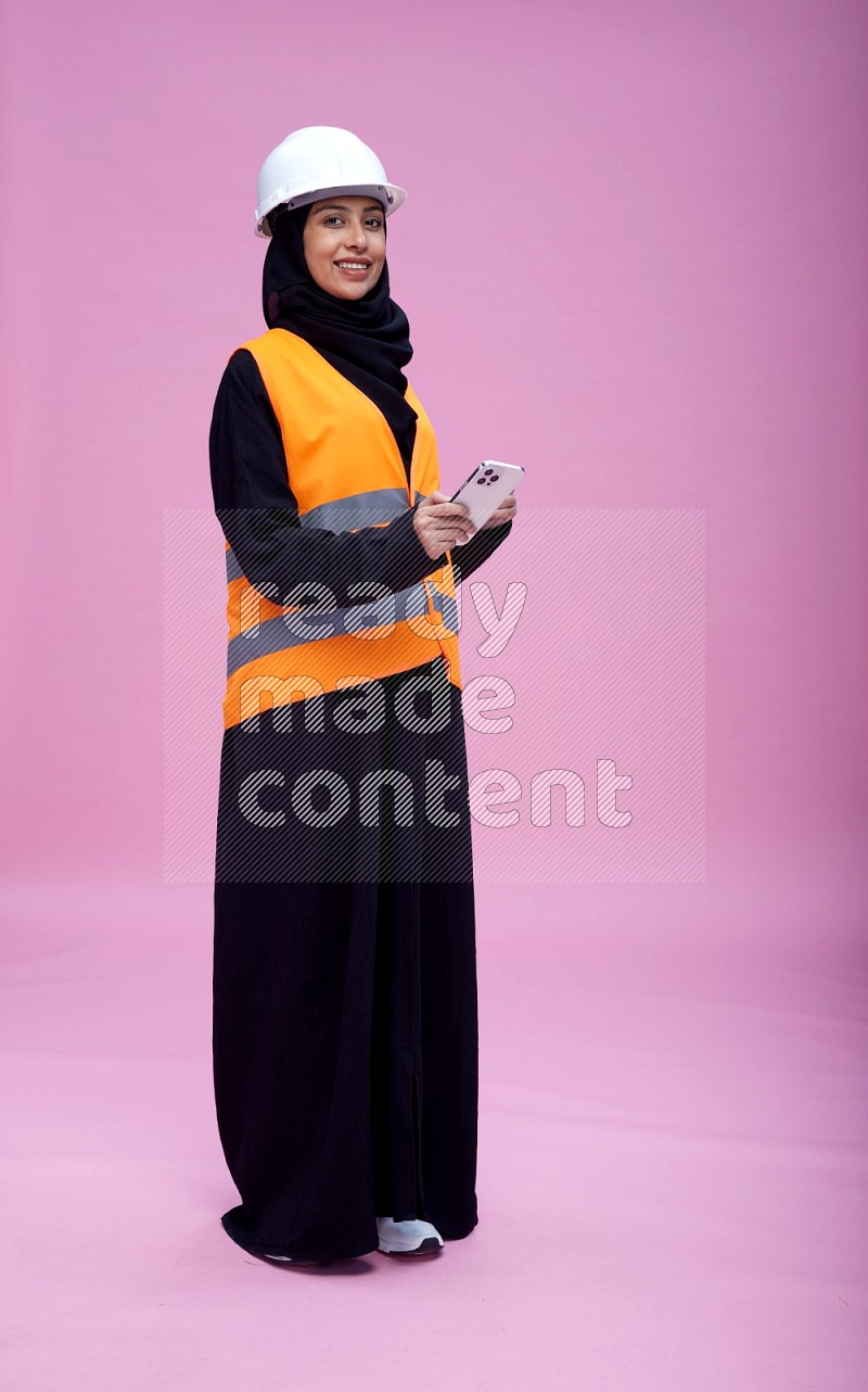 Saudi woman wearing Abaya with engineer vest and helmet standing texting on phone on pink background