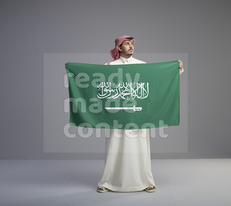 A saudi man standing wearing thob and red shomag holding big saudi flag on gray background