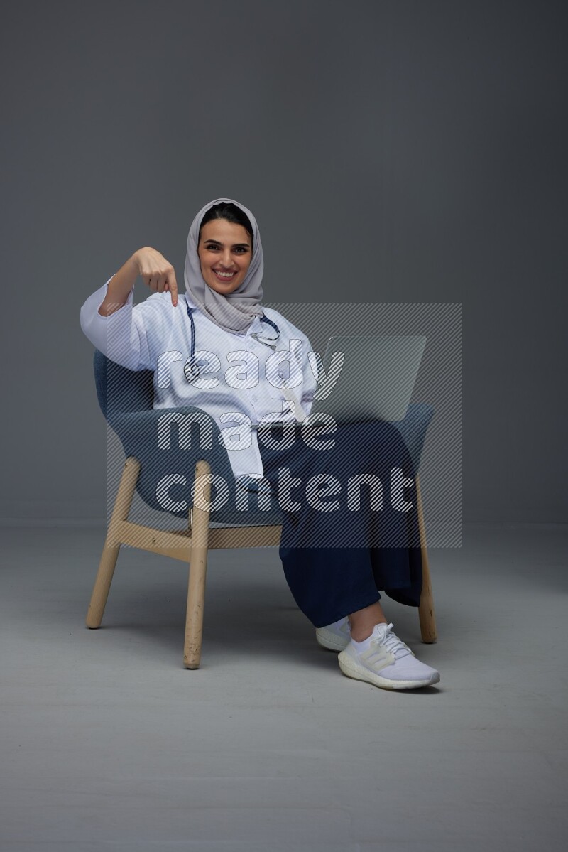 A doctor wearing a light gray head scarf setting on a dark grey chair and using a phone eye level on a grey background