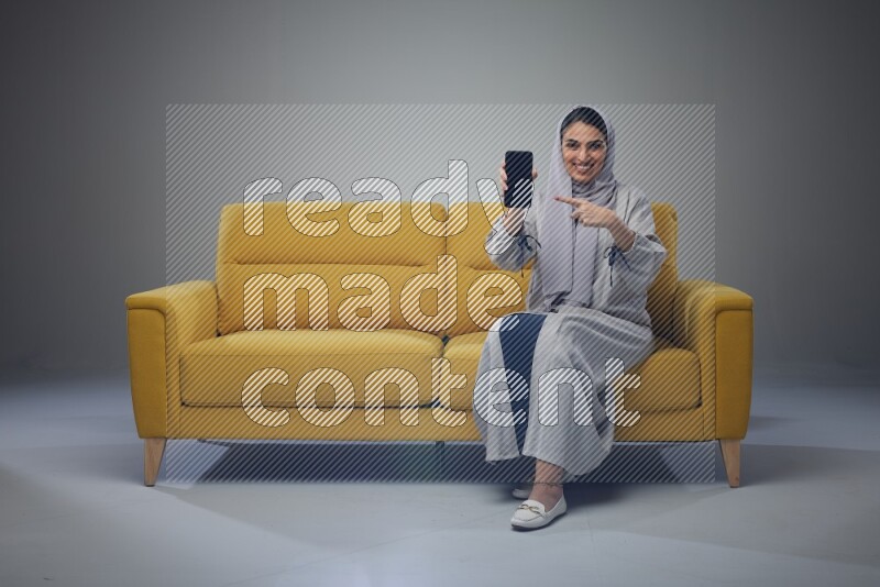 A Saudi woman wearing a light gray Abaya and head scarf setting on a yellow sofa and holding her phone while showing it's screen eye level on a grey background