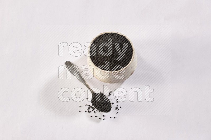 A beige pottery bowl and a metal spoon full of black seeds and more seeds spread on a white flooring