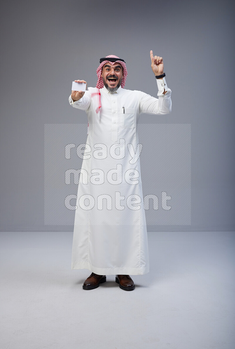 Saudi man Wearing Thob and red Shomag standing holding ATM card on Gray background