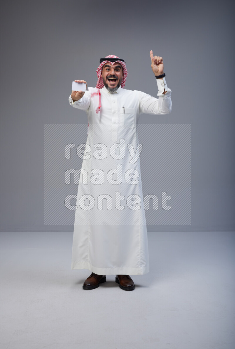 Saudi man Wearing Thob and red Shomag standing holding ATM card on Gray background