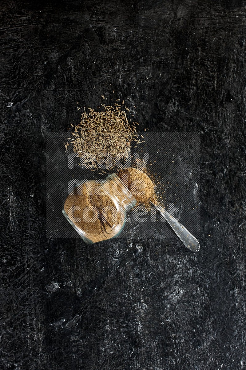 A flipped glass spice jar and a metal spoon full of cumin powder and powder spilled out with cumin seeds on a textured black flooring