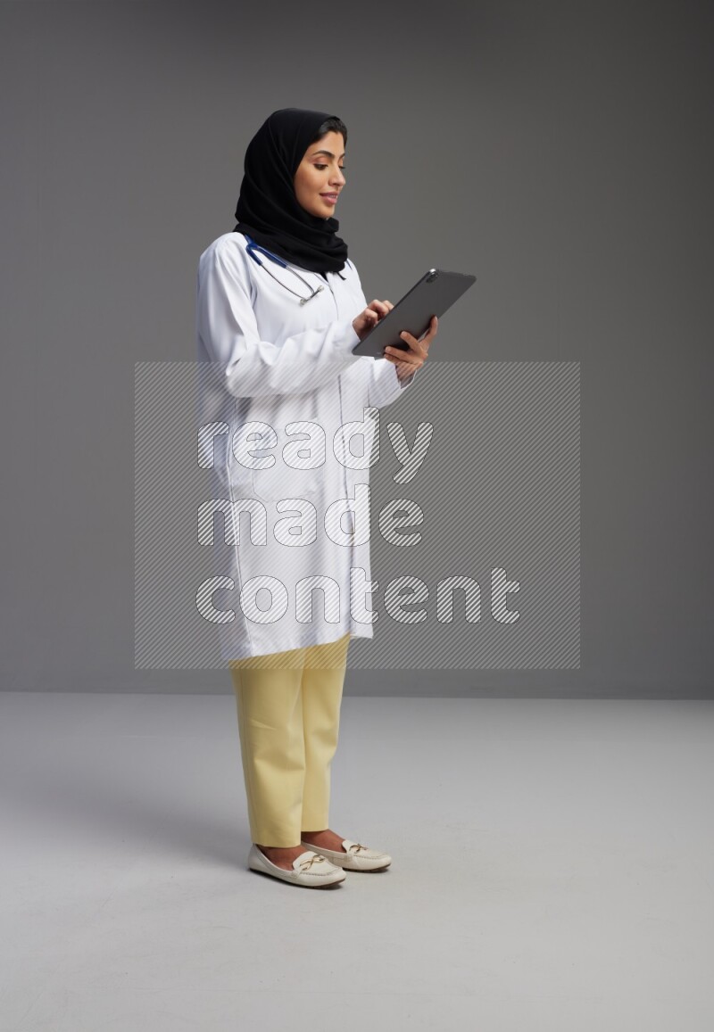 Saudi woman wearing lab coat with stethoscope standing working on tablet on Gray background