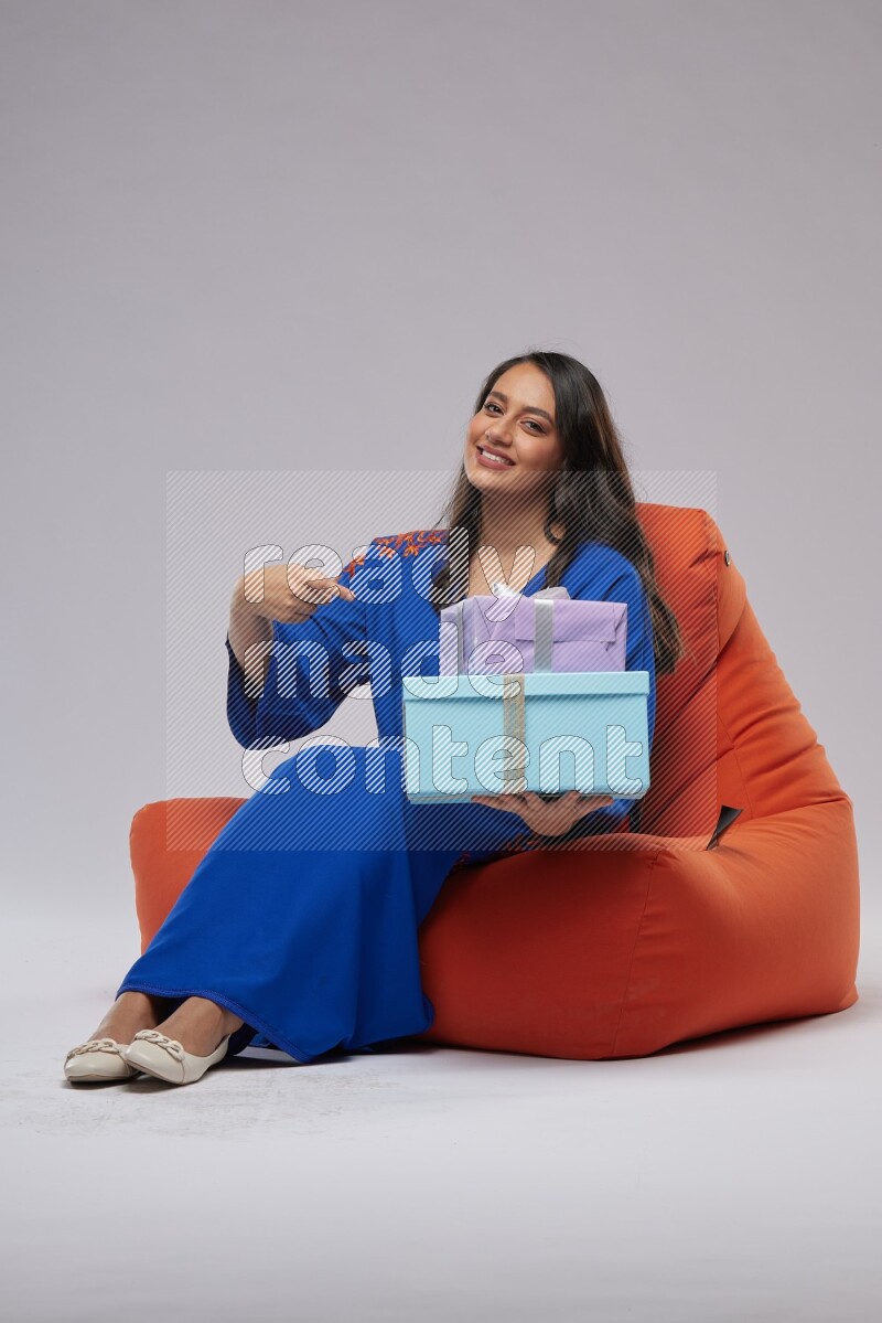 A woman sitting on an orange beanbag wearing Jalabeya holding a gift box